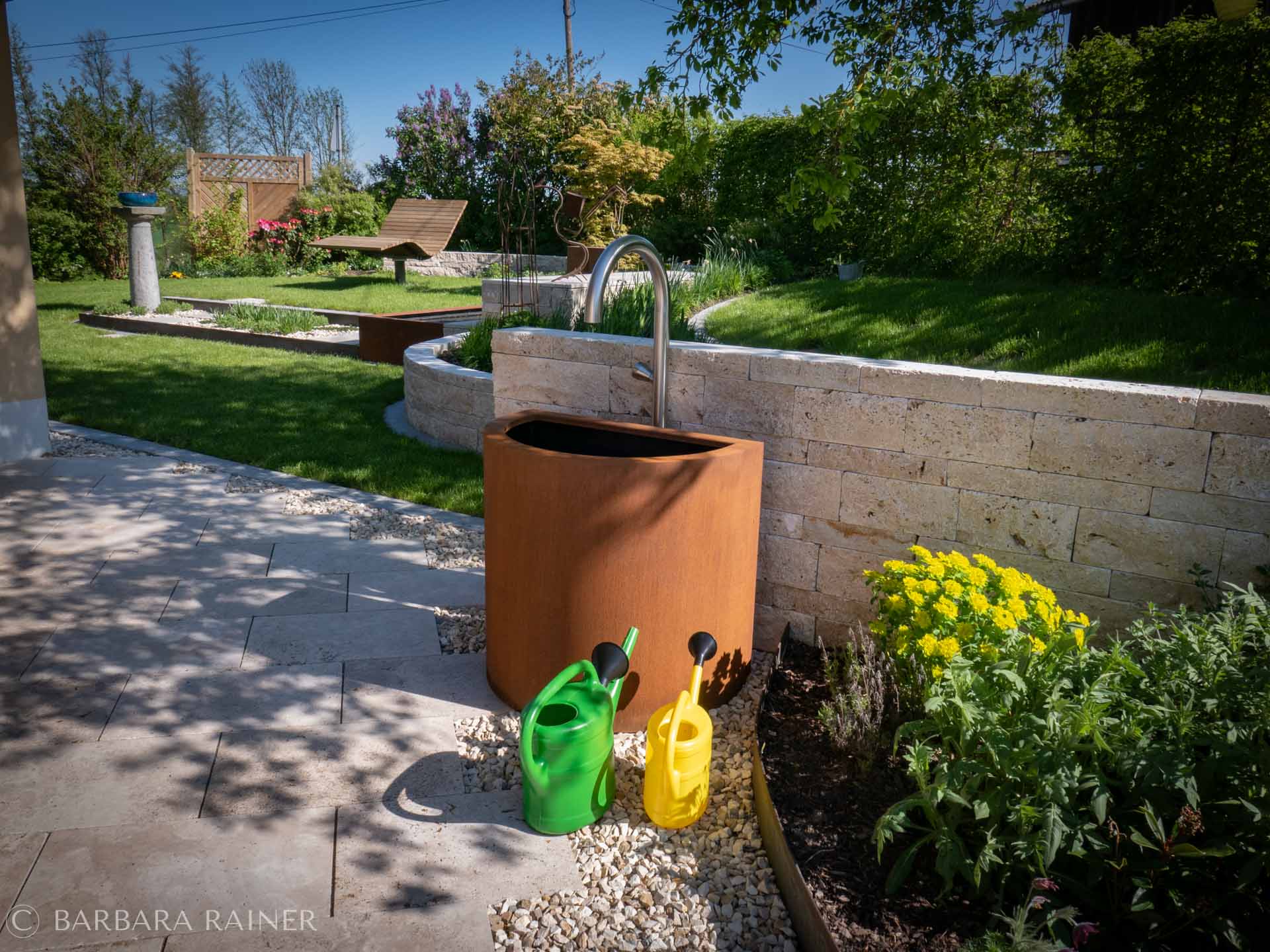 Cortenstahl-Brunnen an geschwungener Travertinmauer mit Wolfsmilch
