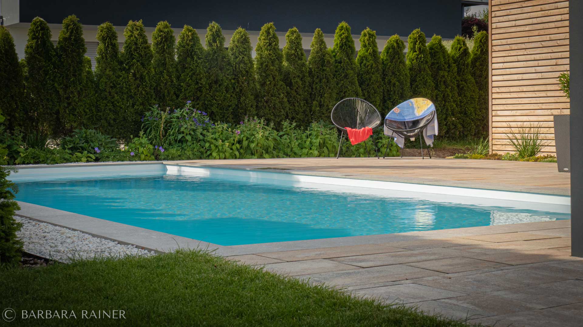 Poolgarten mit Holzdeck, Stauden und Thujen-Reihe im Abendlicht